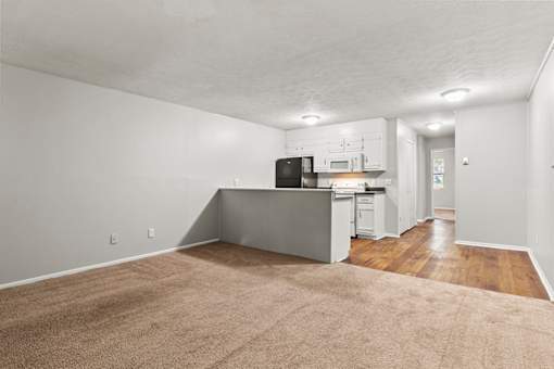 A kitchen with white cabinets and a black fridge is in the background of a living room with a beige carpet.