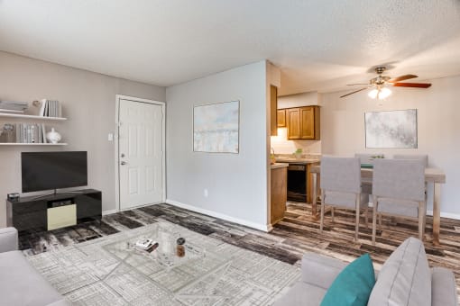 Fort Worth TX Apartments for Rent - Monarch Pass - Living Room with Wood-Style Floor, Beige Walls, Gray Couches, Coffee Table with Clear Glass Top, and a View of the Dining Area and Kitchen