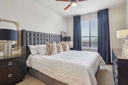 A bedroom with a large bed, a ceiling fan, and a window with blue curtains.