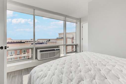 A white bed with a white comforter in a room with a window overlooking a city.
