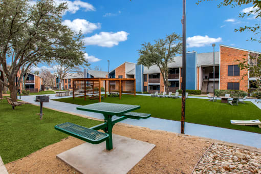 a picnic table sits in the middle of a grassy area with trees and buildings in the