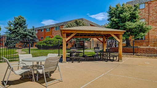 A wooden pavilion with a table and chairs is in the foreground of a sunny outdoor area.