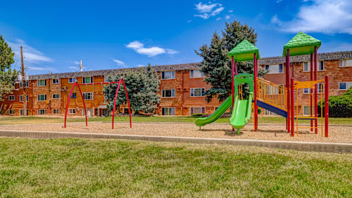 A playground with a green slide and red and yellow play structures in front of a brick building.