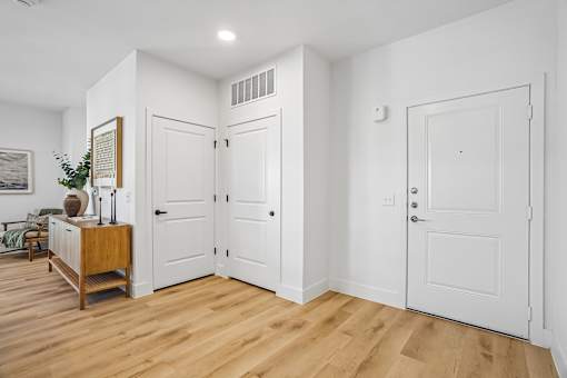 A room with white walls and wooden floors has a white door and a wooden cabinet with a vase on top.