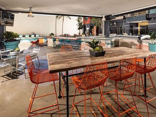 Apartments in Long Beach, CA, with In-Unit Washer and Dryer - Urban Village - Outdoor Kitchen with a Wooden Dining Table, Orange Barstools, and a Small Plant Centerpiece.