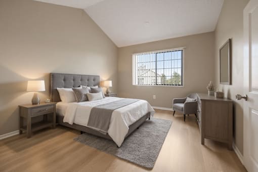 Aurora, CO, Apartments Near Denver Zoo - The Juliet at Lowry - Bedroom with Plank Flooring, Bed, Nightstands, Lamps, Dresser, Armchair, Wall Frame, and a Large Window with Blinds.