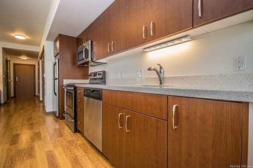 Portland, OR, Apartments for Rent - Corbett Heights - Chef-Inspired Kitchen with Hardwood-Style Floor, Quartz Countertops, Wood-Tone Cabinetry, Stainless Steel Appliances, and View of Hallway.