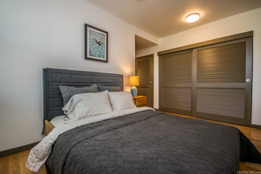 Apartments for Rent in Portland, OR - Corbett Heights - Furnished Bedroom with Hardwood-Style Floor, Large Bed, White Walls, Wooden Nightstand with Lamp, and Spacious Sliding Door Closet.