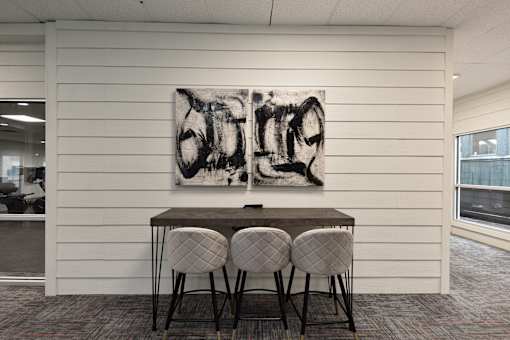 A black and white abstract painting hangs on a white wall above a dark wooden table with four grey chairs.