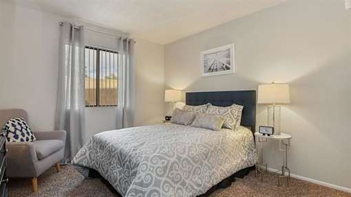 Apartments in Mesa, AZ - Mesa Station - Bedroom with Large Bed, Window with Curtains, Round Bedside Tables, ,Armchair and Carpeted Flooring.