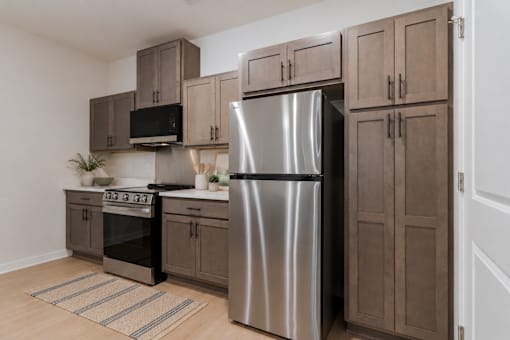Modern kitchen at Blue Sky Landing featuring stainless steel appliances, wood-style cabinetry, efficient appliances, built-in microwave, dishwasher, and ample cabinet space in a brand-new College Park apartment.