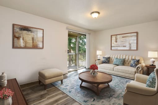 Lynnwood, WA, Apartments for Rent - Evergreen Grove - Living Room with Wood-Style Floor, Area Rug, Couch, Armchair, Stool, Round Coffee Table, Side Tables with Lamps, and Sliding Glass Door with Access to Patio.