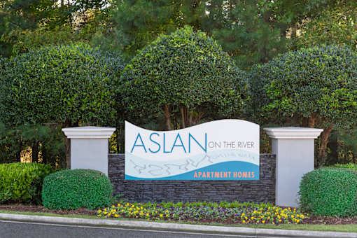 A sign for Aslan on the River Apartment Homes is displayed in front of a green hedge.