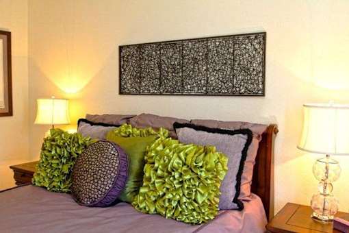 A bed with purple sheets and green pillows is in front of a lamp and a picture.