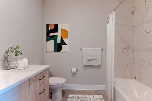 Modern bathroom at Creekline at Fairfield Apartments in Fairfield, CT featuring a soaking tub, marble-style tile surround, wood vanity, quartz countertop, and contemporary finishes.
