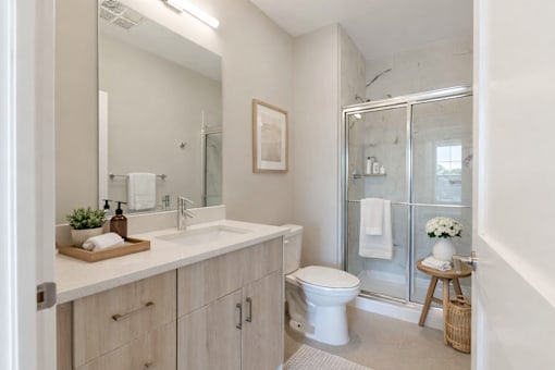 Modern apartment bathroom at Creekline at Fairfield in Fairfield, CT with quartz-style vanity, light wood cabinetry, large mirror, and sliding glass shower with marble-look tile.