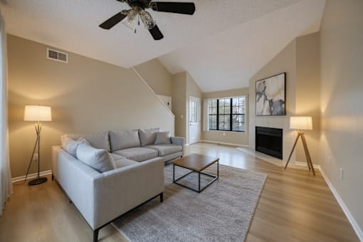 Aurora, CO, Apartments Near Ball Arena - The Juliet at Lowry - Living Room with Plank Flooring, Sectional Couch, Coffee Table, Floor Lamps, Ceiling Fan, Fireplace, Wall Art, Windows, and a Staircase.