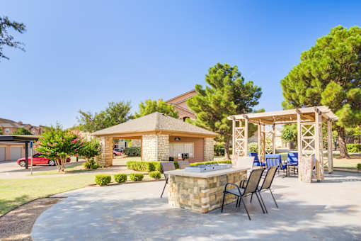 A patio with a barbecue grill and chairs is surrounded by a stone wall and trees.