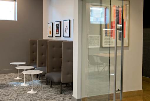 A glass wall separates a room with grey chairs and tables from another room.