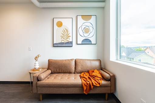 A tan couch with an orange throw is in front of a window with two framed pictures on the wall.