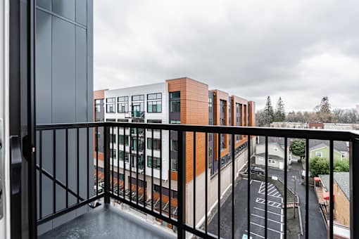 A balcony with a black railing overlooks a parking lot and buildings.
