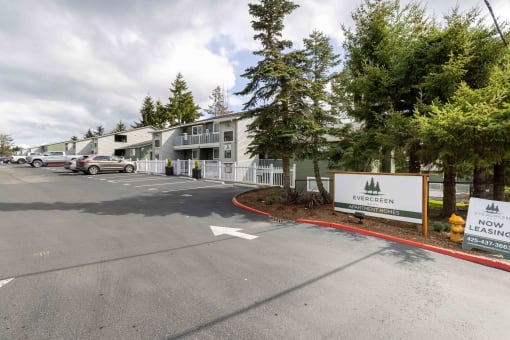 Lynnwood, WA, Apartments - Evergreen Grove - Parking Lot with Vehicles, Trees, Fence, Apartment Buildings, and Property Signage.