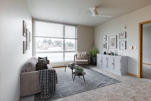 Two-Bedroom Apartments in South Seattle, WA - Greenhouse - Furnished Living Room with Carpeted Flooring, Area Rug, Grey Couch, Armchair, Coffee Table, Grey Cabinet, Ceiling Fan, House Plants, and Extra-Large Window.