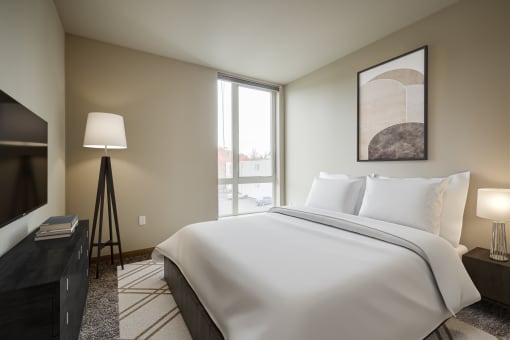 One-Bedroom Apartments in South Seattle, WA - Greenhouse - Bedroom with Carpeted Flooring, Bed, Wooden Nightstand with Lamp, Standing Lamp, Wooden Dresser, Mounted TV, and Extra-Large Window.