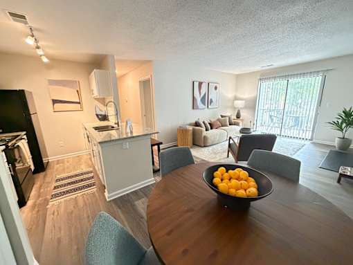 A kitchen area with a refrigerator, a dining table with chairs, and a bowl of oranges on the table.