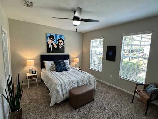 A bedroom with a bed, a ceiling fan, and a painting of two people.
