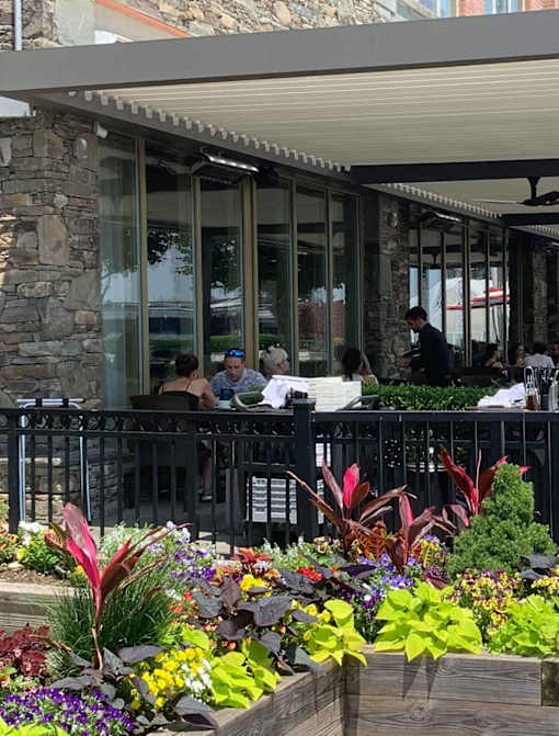 People are sitting at a table outside a restaurant with a stone wall and a wooden planter with colorful flowers.