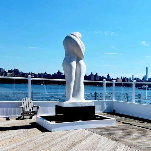 A sculpture of a nude woman stands on a dock with a chair and a cityscape in the background.