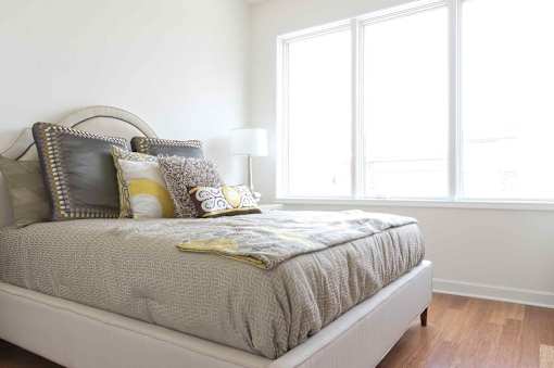 A bed with a grey comforter and a yellow pillow is in front of a window.