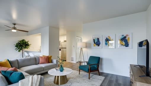 Dog-Friendly Apartments in Everett, WA - Woodbrook - Living Room With A Gray Sectional Sofa, Colorful Accent Pillows, Teal Chair, Round Coffee Table, and Wall Art.