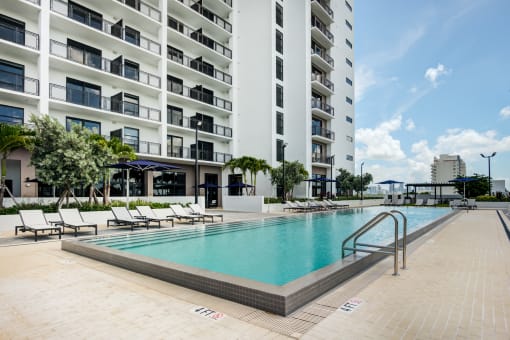 Miami, FL Apartments - MB Station - Apartment Balconies Overlooking Sparkling Resort-Style Pool Surrounded by Lounge Seating and Umbrellas