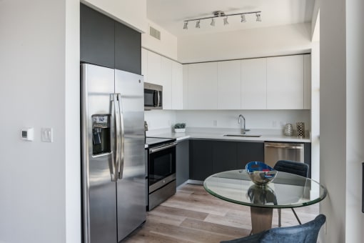Miami Apartments - MB Station - Modern Kitchen With Wood-Style Flooring, Stainless Steel Appliances, White Cabinets, and Round Glass-Top Table with Two Chairs