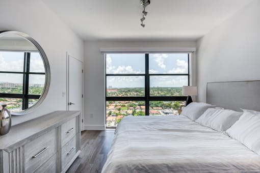 Dog-Friendly Apartments In Miami, FL - MB Station - Spacious Bedroom With Wood-Style Flooring, Modern Light Fixtures, And A Large Window Overlooking The Neighborhood