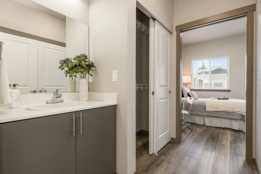 a bathroom with gray cabinets and white countertops and a bedroom in the background,