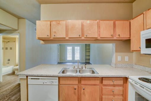 A living room and kitchen with wood flooring and wood cabinets with white dishwasher and white microwave and stove top with oven and steel sink