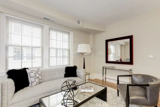 A living room with a grey couch, a glass table, a black leather chair, and a mirror on the wall.
