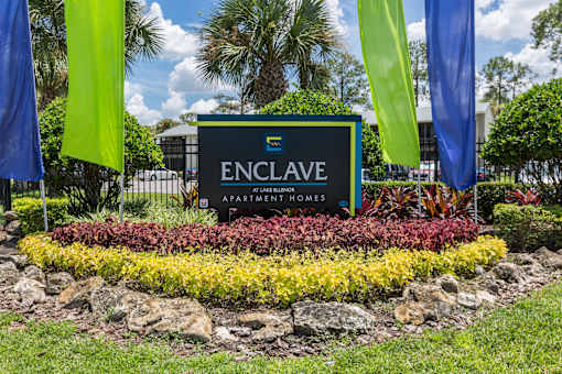 A sign for Enclave Apartments Homes is surrounded by flowers and flags.