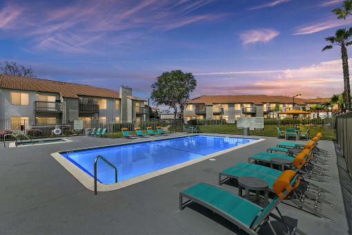 Apartments for Rent in Moreno Valley, CA - Ardella on Chagall - Dusk View of Gated Swimming Pool with Spa, Lounge Seats, Tables and Chairs with Umbrellas, and Apartment Buildings in the Background.