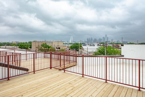A balcony with a red railing and wooden floor overlooks a city skyline.