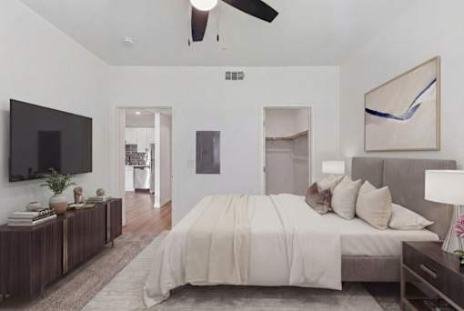 Elegant bedroom showcasing a plush bed with neutral-colored bedding, contemporary decor, and a wall-mounted television, opening into a sleek kitchen area.