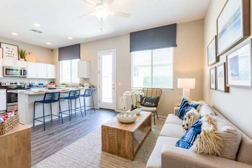 Phoenix, AZ, Apartments near Westgate - The Crofton - Open-Concept Living Room and Kitchen with Breakfast Bar Seating, Blue Stools, Cozy Sofa, Wood Coffee Table, and Large Windows with Soft Light.