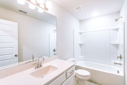 Apartments in Gallatin, TN, for Rent - The Gardens at Preston Park - White Bathroom with Full Tub and Shower, Built-In Shelves, White Cabinetry, Vanity with Quartz Countertop and Sink, Mounted Mirror, and Bright Lighting.