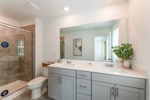 Modern townhome bathroom with double vanity, large mirror, gray cabinetry, and glass-enclosed shower at Townes at Olde Birch in Raleigh, NC.