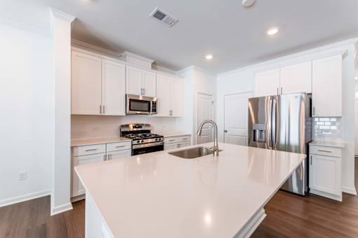 Modern townhome kitchen with oversized island, stainless-steel appliances, white cabinetry, quartz countertops, and wood-style flooring at Townes at Olde Birch in Raleigh, NC.