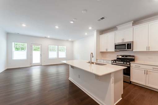 Open-concept townhome kitchen and living area with oversized island, stainless-steel appliances, white cabinetry, wood-style flooring, and private patio access at Townes at Olde Birch in Raleigh, NC.