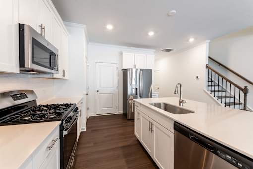 Modern 3-bedroom townhome kitchen with white cabinetry, stainless-steel appliances, quartz island, dishwasher, and staircase view at Townes at Olde Birch in Raleigh, NC.